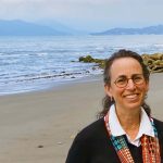 A photo of Dr. Bonnie Effros in Vancouver. She is on a sandy beach with large boulders on the shore. Mountains are visible behind her. She has brished back dark brown hair and wears round glasses.
