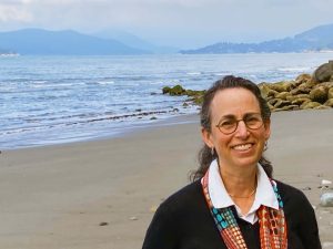 A photo of Dr. Bonnie Effros in Vancouver. She is on a sandy beach with large boulders on the shore. Mountains are visible behind her. She has brished back dark brown hair and wears round glasses.