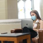 Stock image of a person sitting in a brown armchair. They are holding a laptop, and there is a small coffee table in front of their legs. They have long brown hair, and they are wearing a blue medical mask.