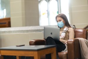 Stock image of a person sitting in a brown armchair. They are holding a laptop, and there is a small coffee table in front of their legs. They have long brown hair, and they are wearing a blue medical mask.