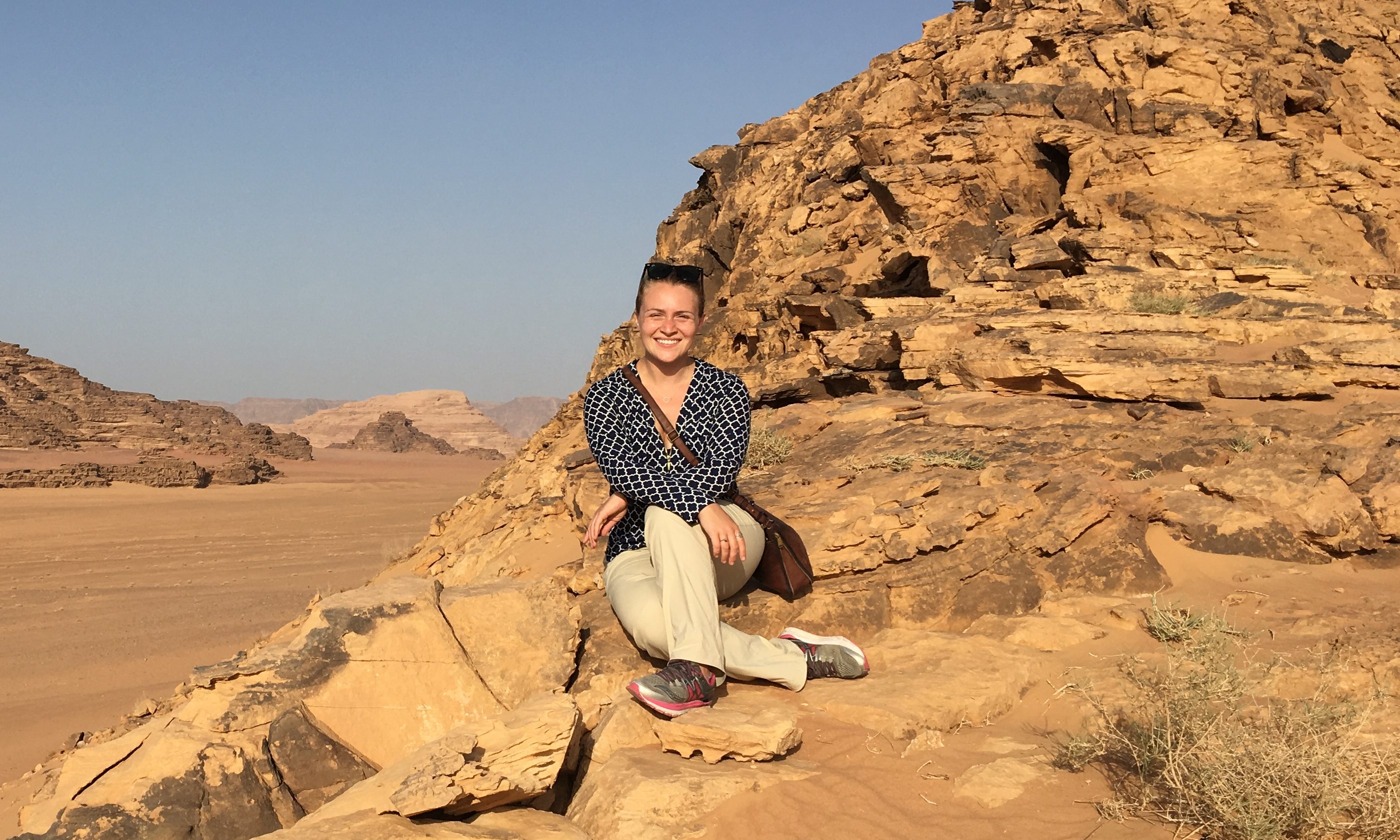 Dr. Sara Ann Knutson in the Wadi Rum, Jordan. She wears a black shirt with white line patterns, khaki pants, and white, silver, and pink running shoes. She smiles and sits on a pile of sand stone with one leg crossed in front of the other.