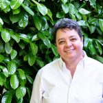 Joy Dixon smiles in front of a tall green bush. They have short salt and pepper hair and wear a white button down shirt. She smiles at the camera.