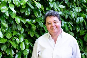 Joy Dixon smiles in front of a tall green bush. They have short salt and pepper hair and wear a white button down shirt. She smiles at the camera.